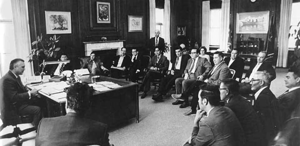 Men in suits sitting around a desk.